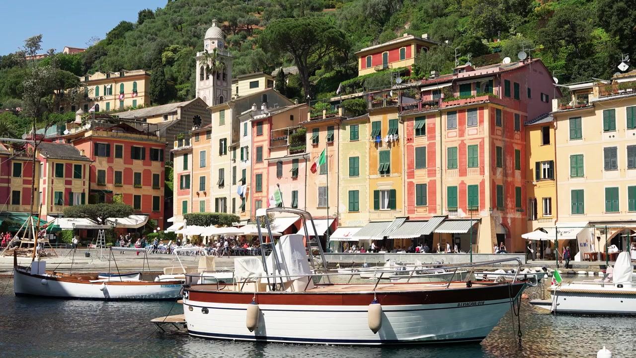 Barche vivaci ormeggiate nel porto di Portofino con edifici storici color pastello, una collina lussureggiante e acqua scintillante sotto la chiara luce del giorno. Composizione luminosa e vivace, nessuna persona visibile.
