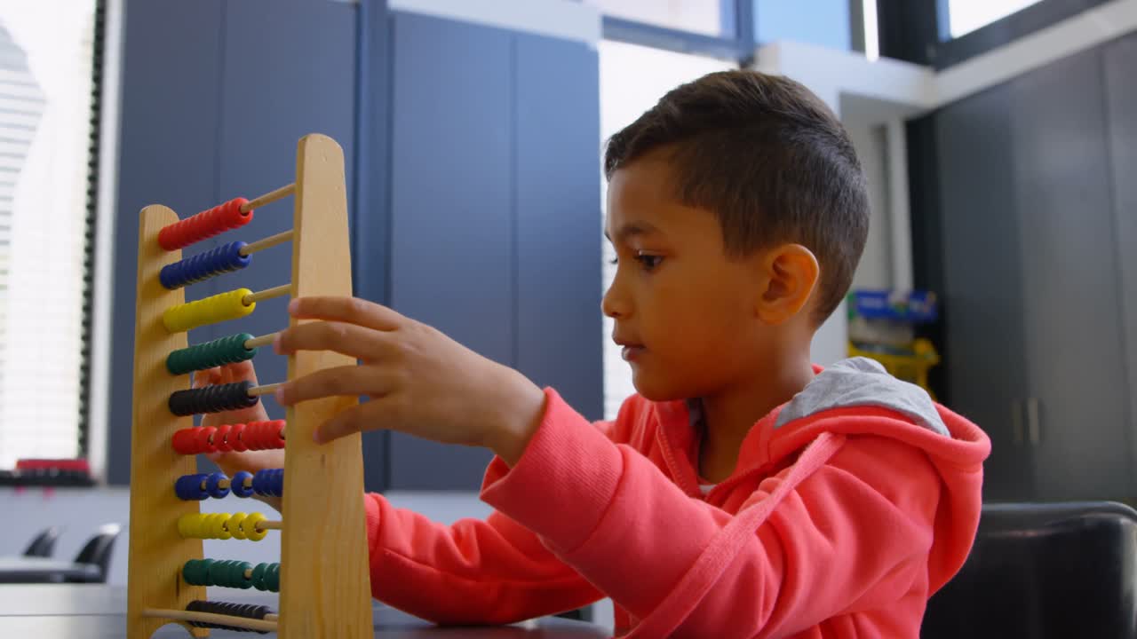 Vista lateral de un niño escolar asiático resolviendo un problema de matemáticas con un ábaco en su pupitre en un aula de la escuela 4k