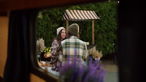 Shooting-from-the-window-of-a-trailer,-a-brunette-girl-in-a-white-hat-and-a-pink-checkered-shirt-communicates-with-her-brunette-boyfriend-in-a-checkered-shirt-who-is-sitting-at-a-table-outside-the-city-in-a-camp-during-their-picnic-in-the-summer