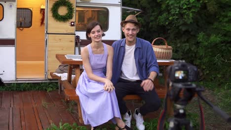 una pareja de bloggers populares grabando videos sobre ellos mismos, sentados en la mesa de madera frente a la casa de las ruedas, hablando y mirando la cámara en un trípode
