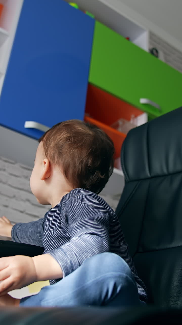 Smiling cute kid sitting in the turning black leather chair. Toddler turns to the table to take remote control. Low angle view. Vertical video