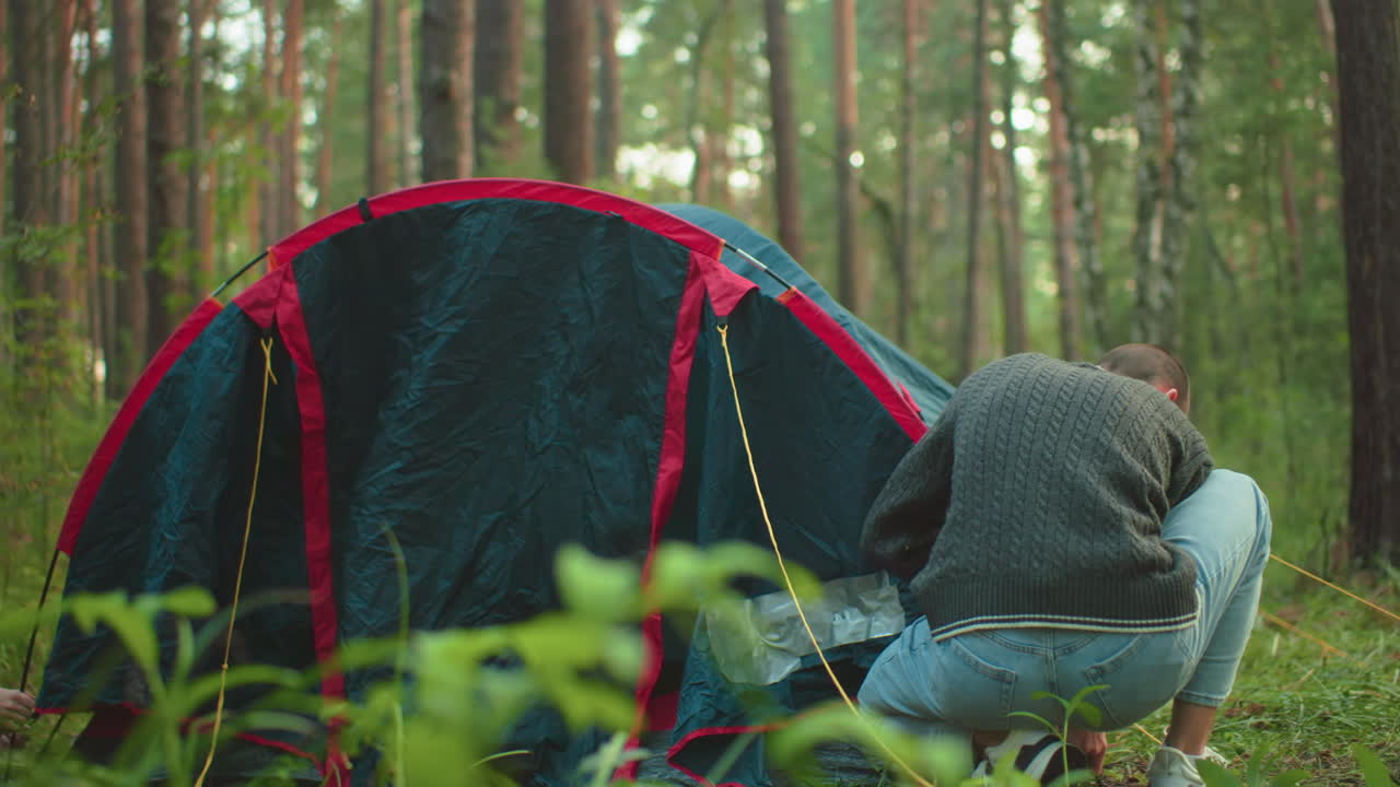 Couples work together assembling camping tent in lush green forest, one crouching to support pole while other secures tent edges, surrounded by trees and natural vegetation