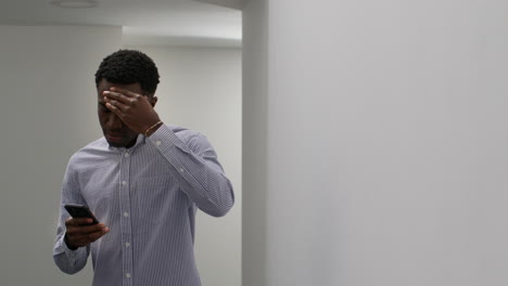 young businessman in office looking at message with bad news on mobile phone and leans against wall looking unhappy and depressed