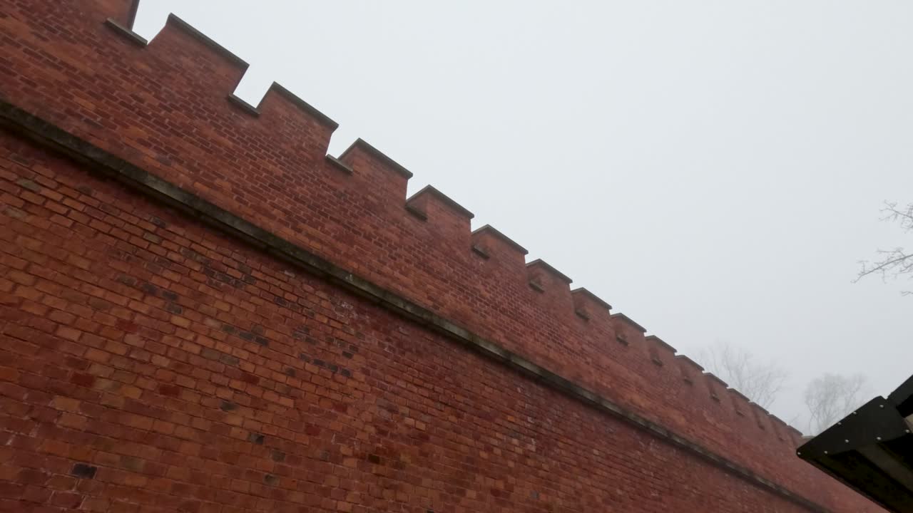 In a chilly morning setting, an old red brick wall stands tall against a gray sky. The structure shows signs of age and history, surrounded by dense fog