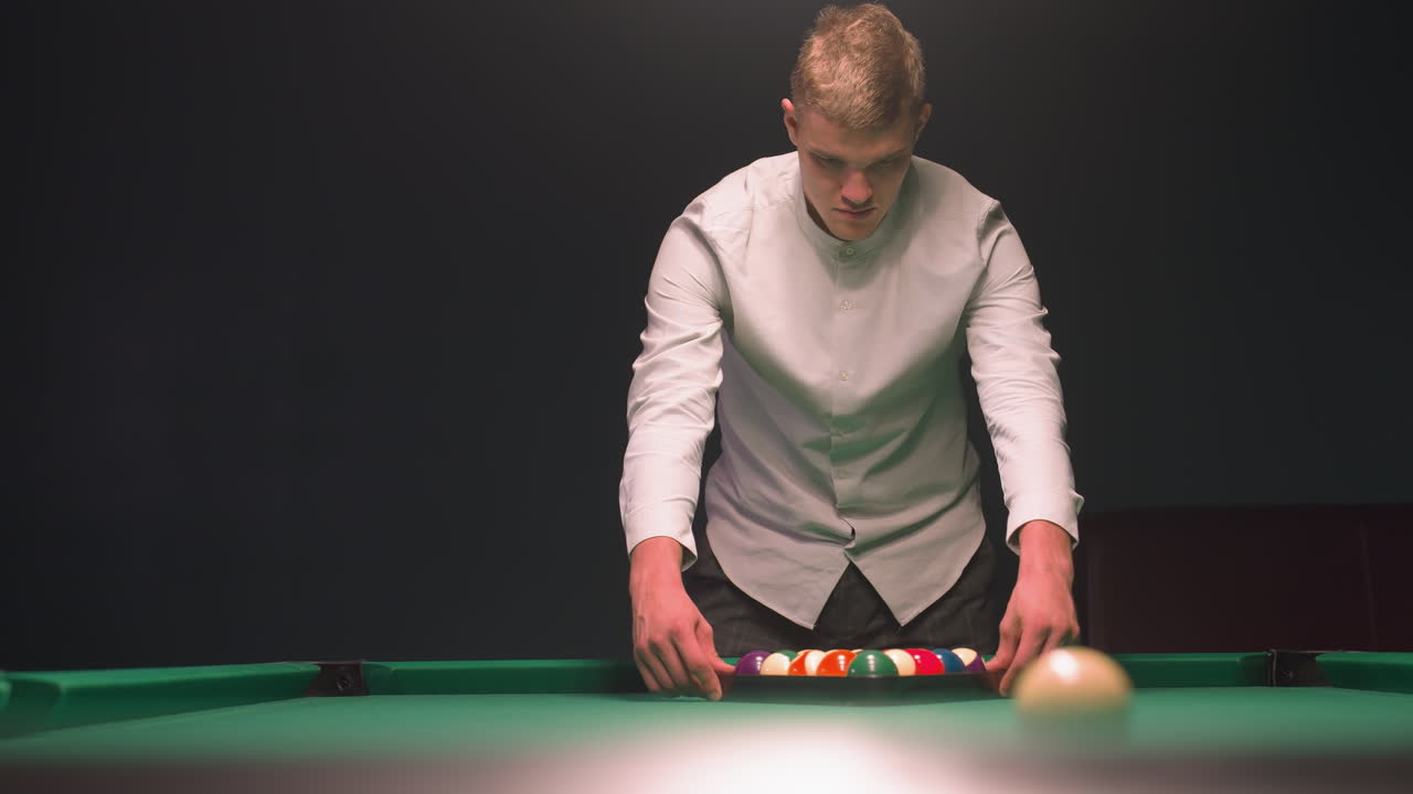 camisa de aprendiz de billar y pantalones grises arregla cuidadosamente bolas de billar de colores en triángulo en la mesa de billar verde. mientras se centra con concentración tranquila en el salón elegante