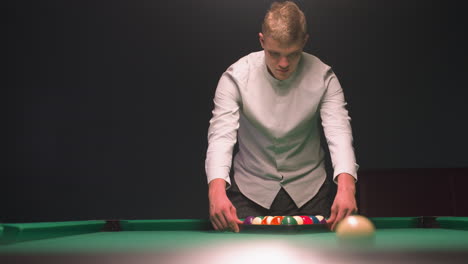 billiards trainee shirt and gray trousers carefully arranges colorful billiard balls in triangle on green pool table. as he focuses with calm concentration in stylish lounge