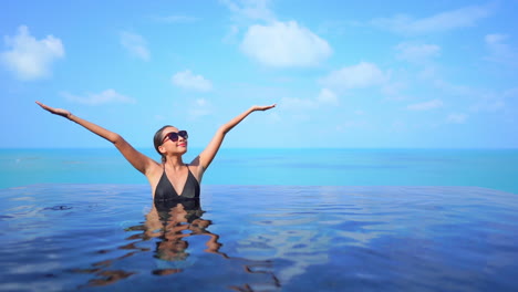 mujer hermosa medio sumergida de pie en la piscina infinita levantando los brazos por encima de la cabeza en un cielo azul