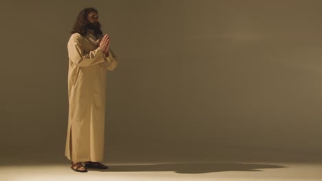 full length studio portrait of man wearing robes and sandals with long hair and beard representing figure of jesus christ praying