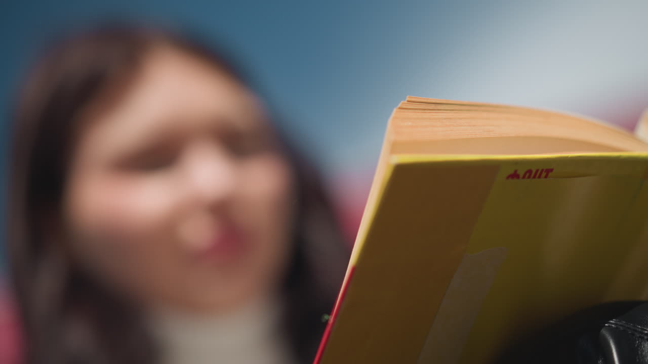 primer plano de una dama en un entorno al aire libre, enfoque borroso en el libro de portada amarilla mientras hojea las páginas, la luz del sol crea un brillo suave, con el fondo suavemente borroso