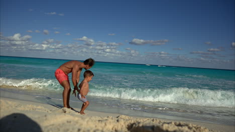 cámara lenta de un joven mexicano latino con pantalones cortos huyendo del agua en la playa riendo y sonriendo divirtiéndose con su padre