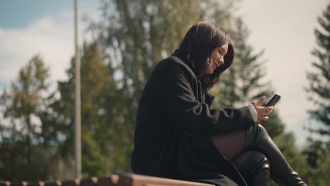 side view of a young woman sitting on a bench in a park, dressed in a black coat and knee-high boots, focusing on her smartphone, she smiles while surrounded by trees and greenery on a sunny day
