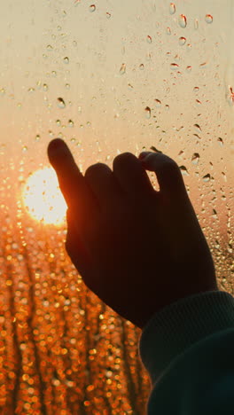 silhouette of finger trying to touch evening sun behind wet window closeup. hand touches glass with leaking water pattern. dreaming at dusk