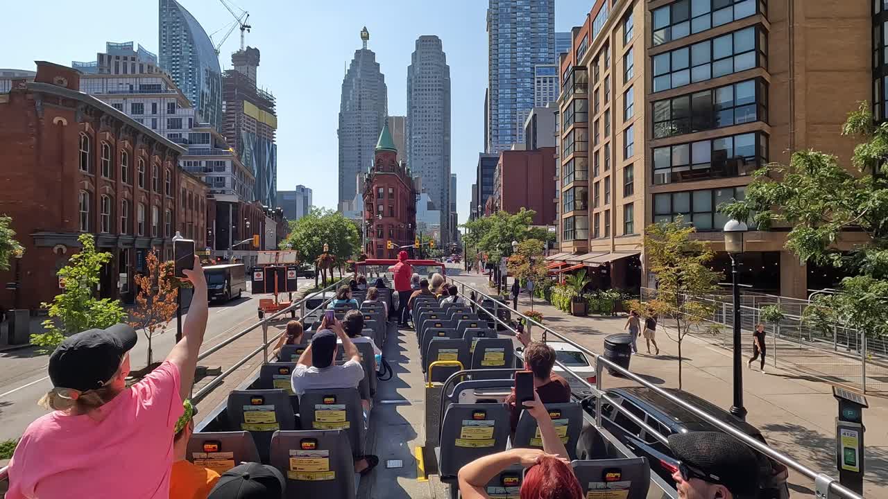 First-person view of Toronto Front Street from moving hop-on hop-off sightseeing bus, with tourists seated enjoying tour among towering skyscrapers and bustling car traffic, Canada