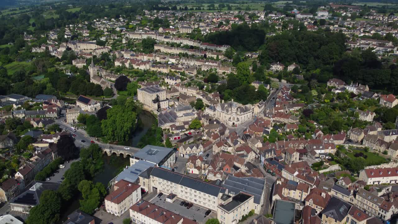Drone rises above the town of Bradford-on-Avon, revealing its stone buildings, bridge, church, and green surroundings before tilting down into a top-down view of the city