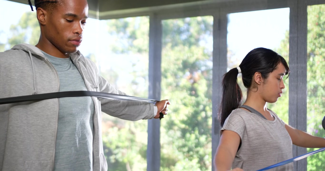 African American coach holding black band, Asian trainee pulling blue handles at gym for strength