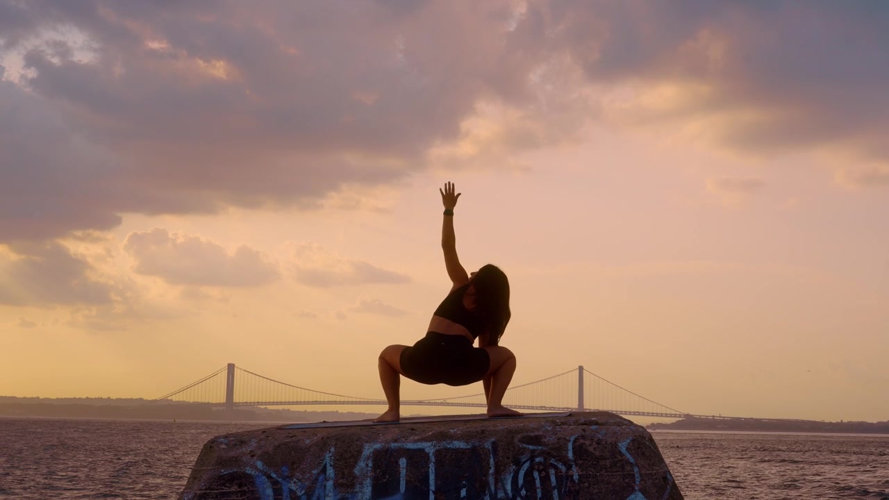 Donna che esegue una posa yoga di squat profondo al tramonto a New York. Postura a gambe larghe sul bordo di pietra con cielo scuro e sfondo del ponte Verrazzano Narrows