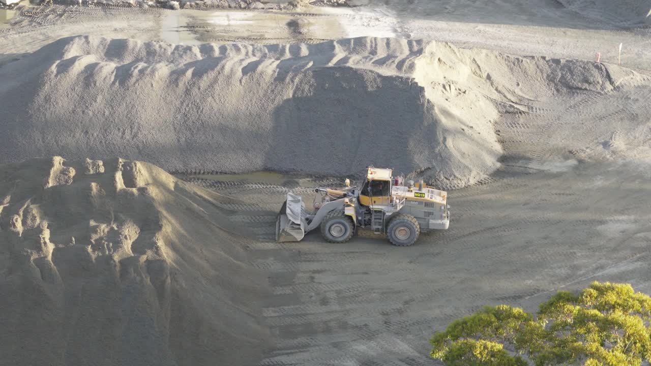 Aerial footage of a loader moving soil at a Gold Coast quarry. Bright daylight highlights the machinery and sandy terrain