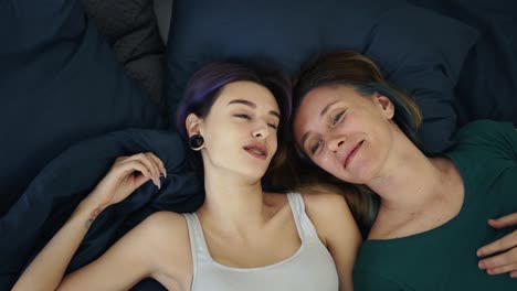 top view of two young women lying in bed, talking to each other and smiling