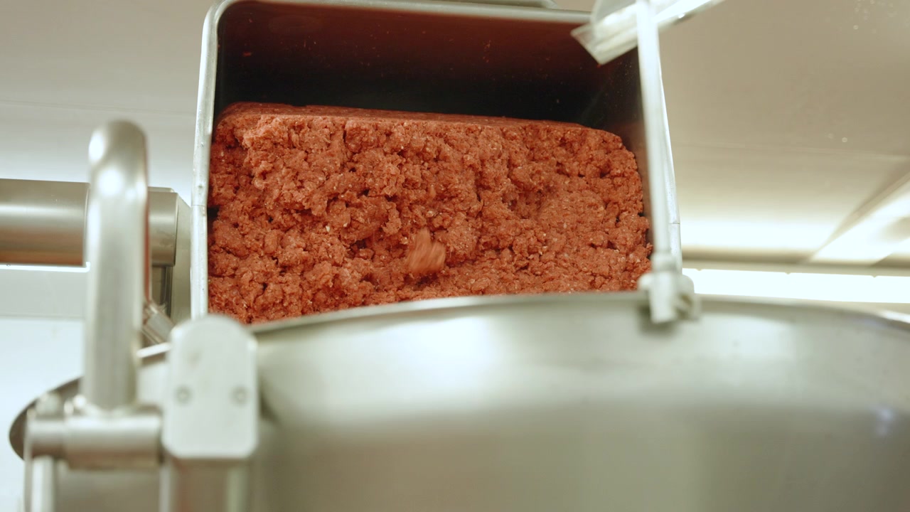 Chunky minced beef falls from a metal hopper into a large industrial mixer inside a modern meat processing line.