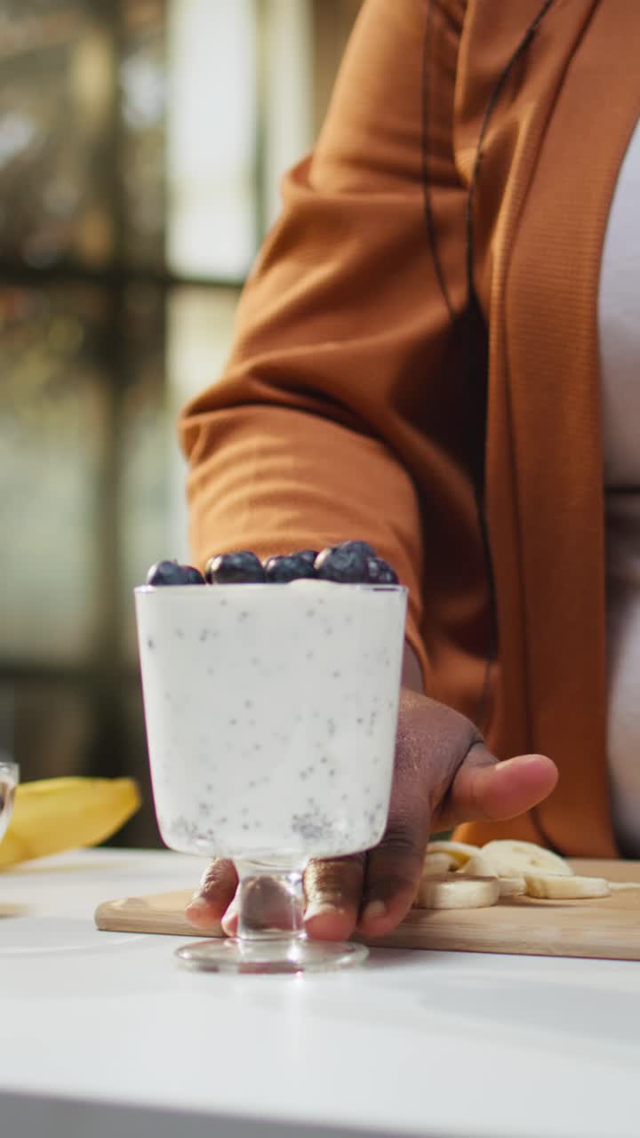 Video verticale. Influencer donna di colore che aggiunge mirtilli su una ciotola di yogurt e semi di chia.