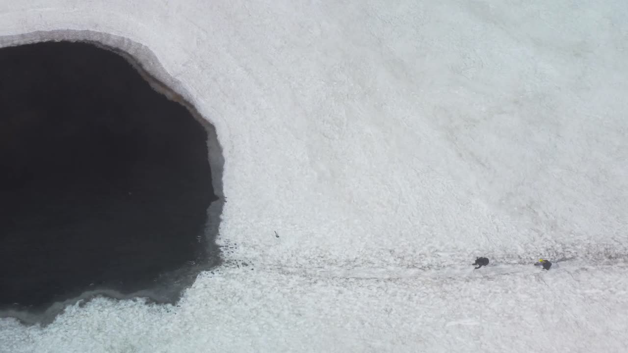 Top down aerial view of two hikers walking on Iceland snow covered dark volcanic landscape and approach a dark, cold and icy lake in the nature. People create footsteps in snow while walking forward.