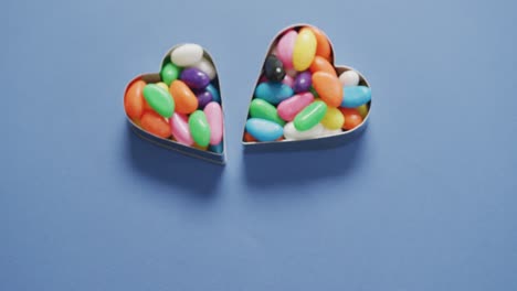 Video-of-overhead-view-of-multi-coloured-sweets-in-two-heart-shapes-over-blue-background