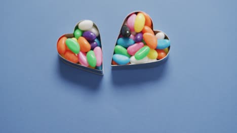 video of overhead view of multi coloured sweets in two heart shapes over blue background