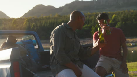 two male friends with backpacks in pick up truck on road trip by lake drinking beer and doing cheers