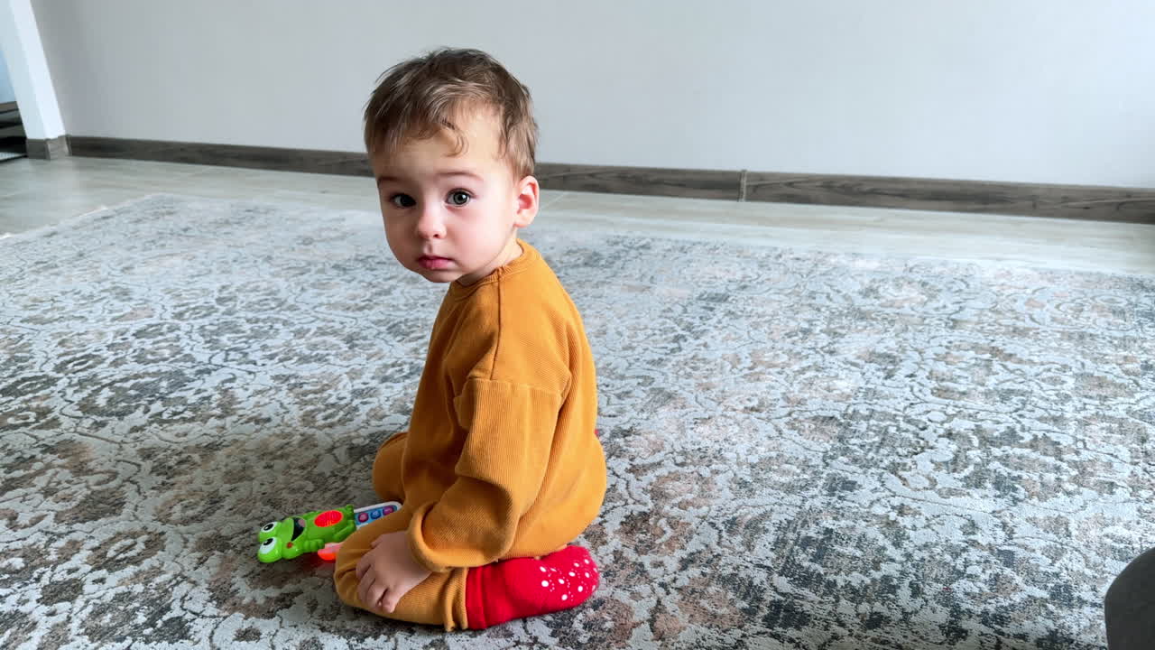 Lovely little kid sits on the floor. Baby boy has a toy phone playing in front of him. Child turns to look at camera.