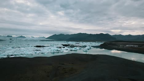 Panoramic-view-of-Iceberg-drifting-on-the-glacier-water-surface,-Melting-glaciers-due-to-global-warming,-Mountains-under-an-overcast-sky,-Iceland,-Slow-motion-drone-shot