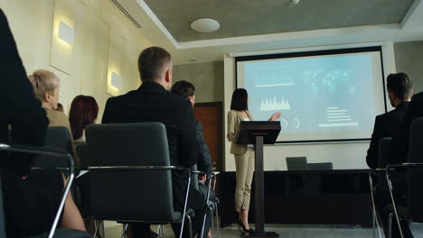 caucasian businesswoman talking on a podium in a conference room and showing some charts and graphics on the big screen