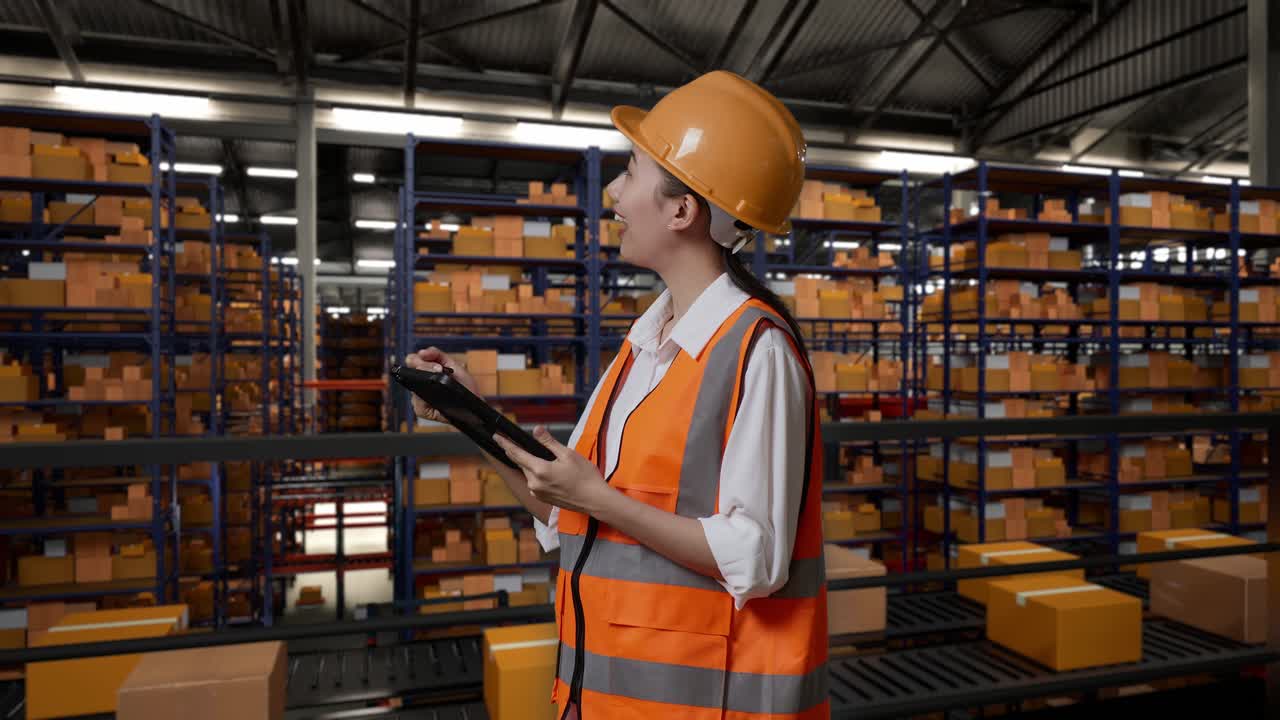 vista lateral de una ingeniera asiática con casco de seguridad de pie en el almacén con estantes llenos de bienes de entrega. tomando nota en la tableta y mirando a su alrededor en el almacenamiento