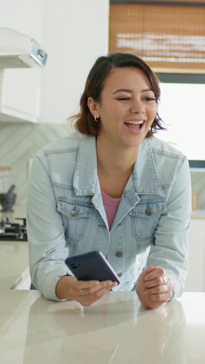 Video verticale: Donna che ride in giacca di jeans, reagisce a una notifica del telefono all'isola della cucina, smartphone.