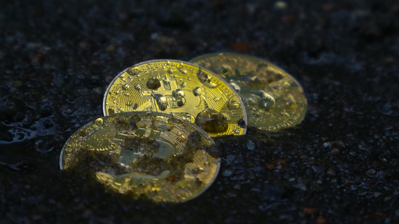 Primo piano di monete Bitcoin dorate che affondano nel fango scuro e bagnato mentre cadono gocce di pioggia, a simboleggiare il crollo del mercato delle criptovalute, la crisi finanziaria o la perdita e il fallimento degli investimenti