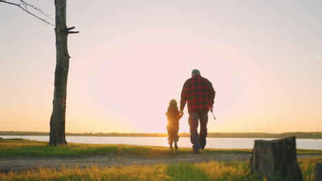 Der-ältere-Fischer-Und-Sein-Kleiner-Enkel-Gehen-Im-Sonnenuntergang-Zum-Seeufer.-Opa-Trägt-Angelrute-Und-Rucksack