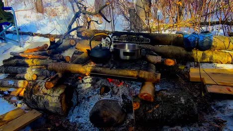 winter campsite fire log cabin style design stack of wood with a forest background snow covered grounds foldable chair metal rack overtop the flames with a tea kettle and a pot steaming boiling sunset