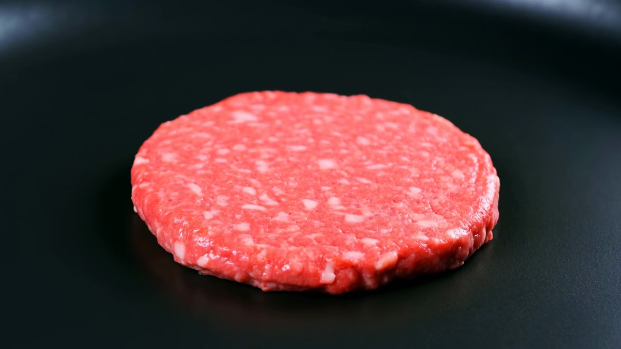 Close-up view of raw ground meat patty on dark flat cooking surface, shown from low angle with blurred background, coarse moist texture, white fat flecks, gentle sheen, fresh minimal culinary mood
