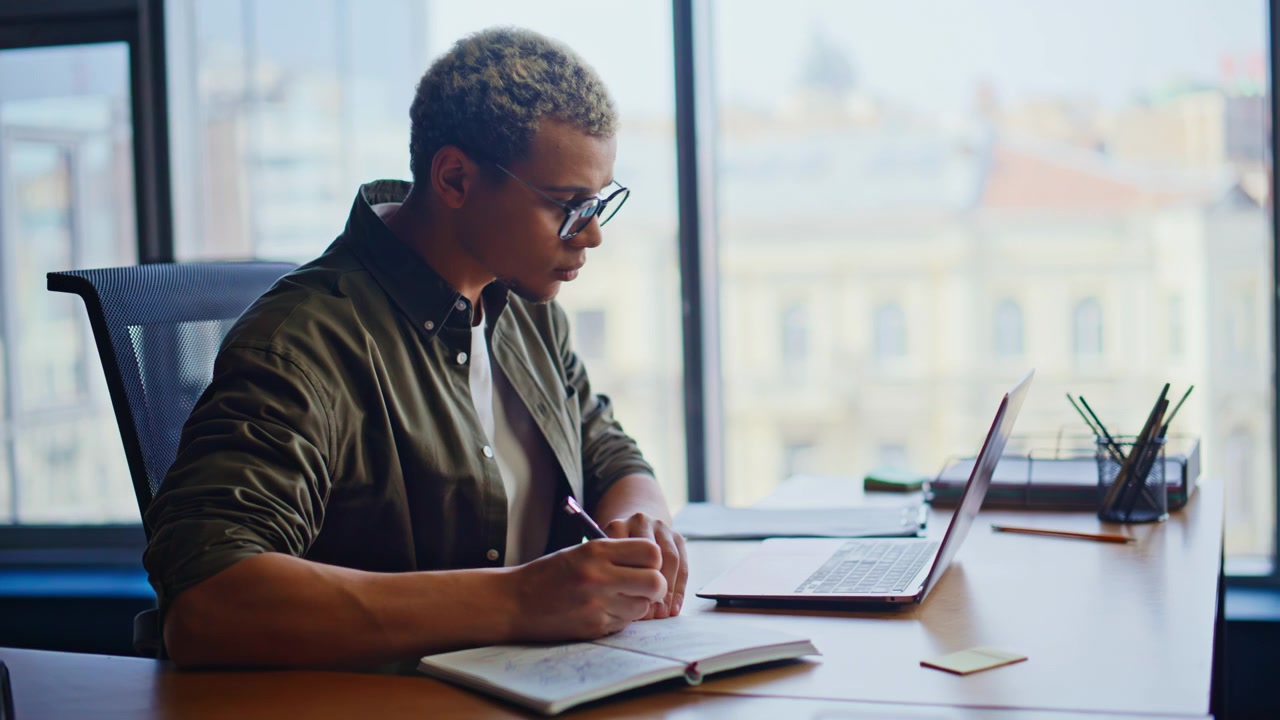 Focused african american man typing laptop office. Businessman writing notes