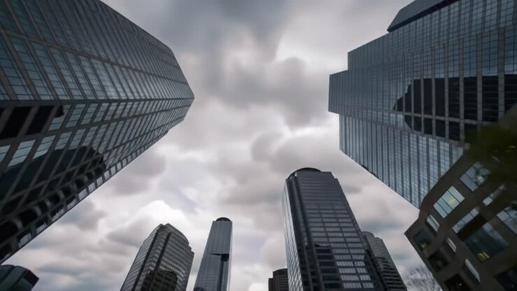 Tall glass skyscrapers stretch towards the cloudy sky, creating a striking urban landscape. The buildings reflect the muted light while the city prepares for possible rain.