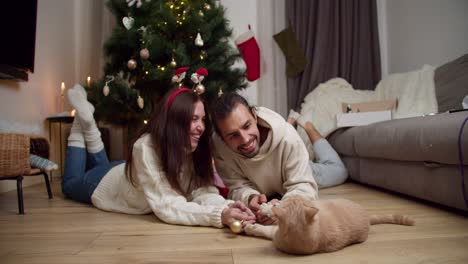 happy couple, a guy and a brunette girl lie on the floor and play with their cream-colored cat with the help of a new year's toy near a decorated new year's tree in their cozy home in winter