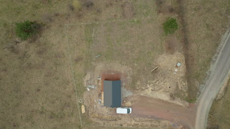 una toma aérea revela un sitio de construcción aislado, con un pequeño edificio y materiales esparcidos, con una única furgoneta blanca estacionada al lado