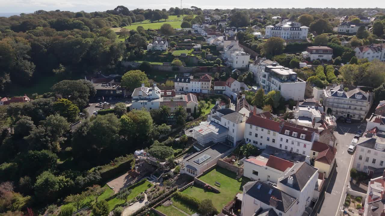 High drone footage over residential area of St Peter Port Guernsey heading towards open fields showing houses and gardens on bright sunny afternoon