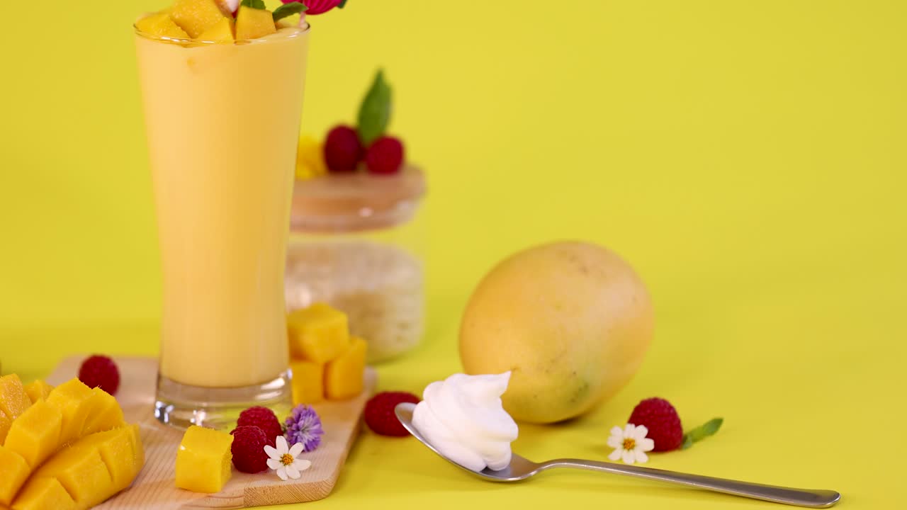 Mango smoothie, cut mango, raspberries, whipped cream, and flowers arranged on bright yellow background