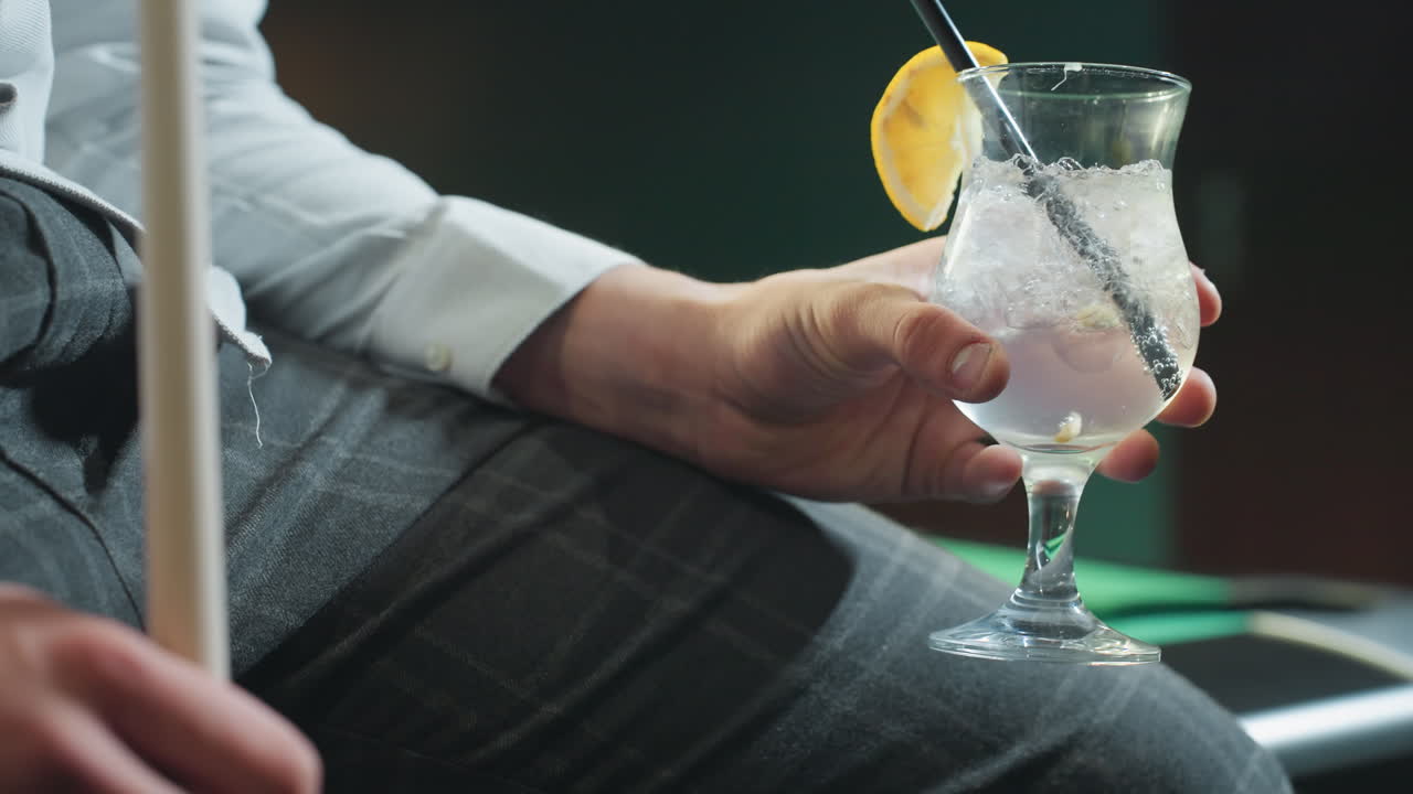 Primo piano di una persona, viso non visibile, che indossa una camicia bianca e pantaloni grigi, tiene in mano una stecca da biliardo e un bicchiere di limonata con cannuccia nera. Cubetti di ghiaccio e fetta di limone esaltano l'atmosfera rinfrescante.