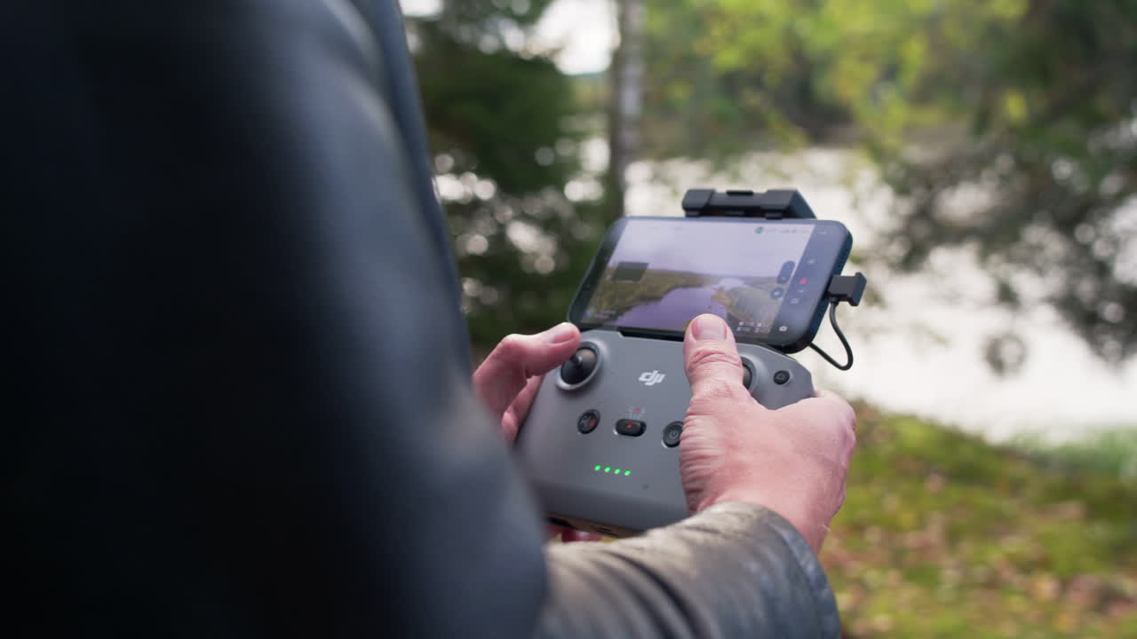 A white man's thumbs operate a DJI controller as he sees the vidoe of the drone on his phone