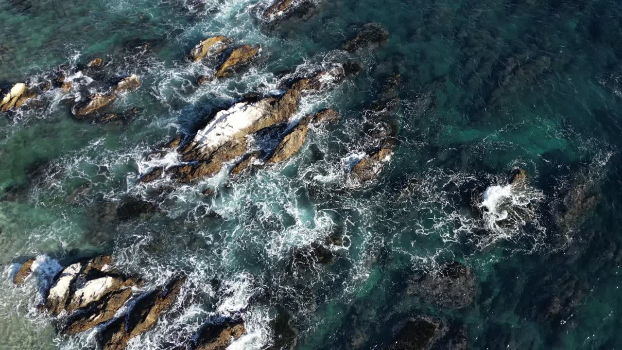 Riprese aeree di onde oceaniche che si infrangono contro una costa rocciosa con acqua limpida e texture naturali. Il potente movimento del mare crea un paesaggio costiero dinamico dall'alto.