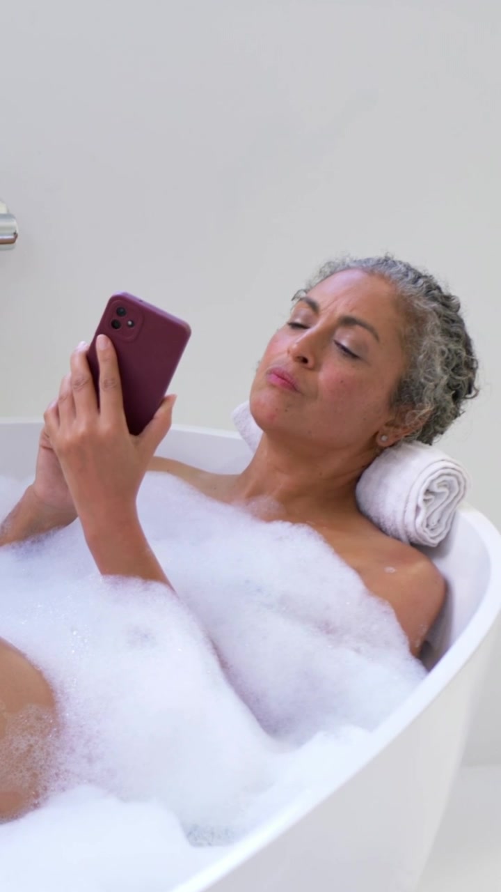 Vertical video: Holding purple phone, woman on towel in tub tapping and frowning in foam, tap