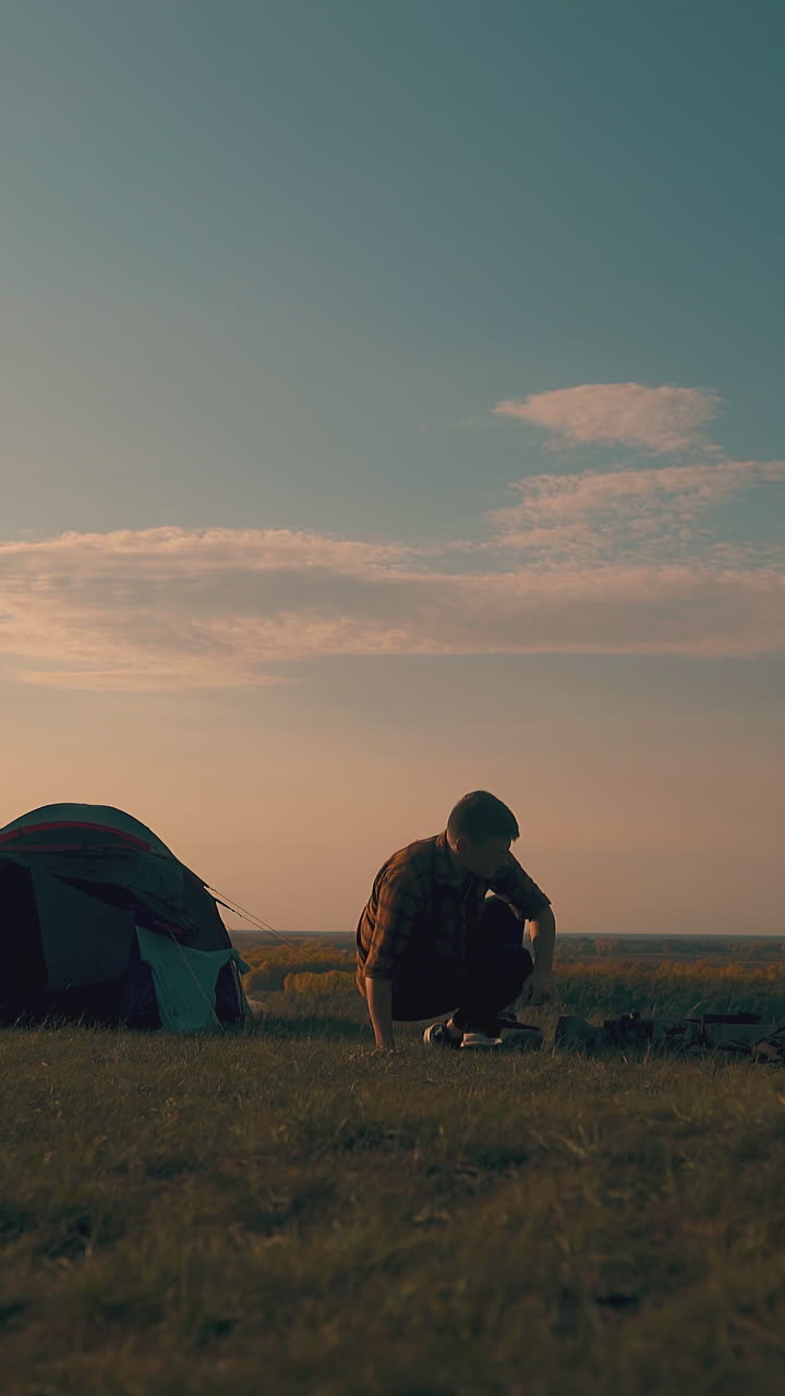 man silhouette comes to bonfire from tent in camp on river bank in cool autumn morning at sunrise
