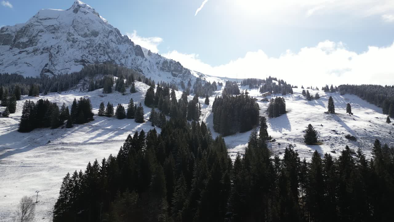 Aerial drone pan shot from left to right over snow covered mountain slope with mountains in the background on a sunny day