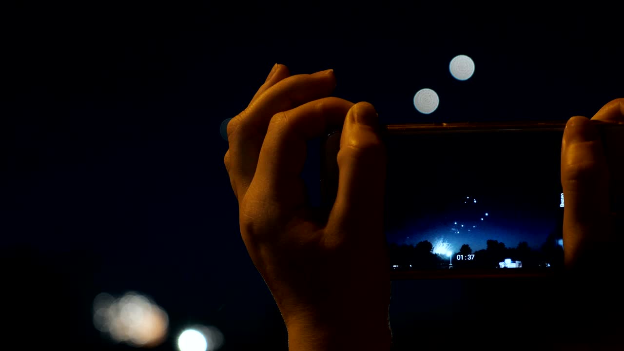 한 여성이 스마트폰으로 불꽃놀이를 니다. 보케 빛의 배경으로 불꽃 놀이를 축하하고 있습니다.